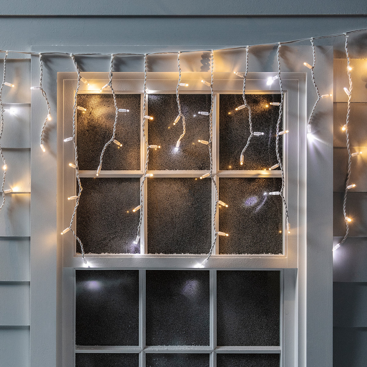 Glitzernde LED Eiszapfen-Lichterkette hängt beleuchtet vor einem Fenster mit weißem Rahmen