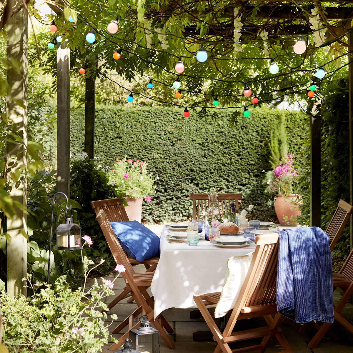 LED-Lichterkette bunt leuchtend unter grünem Laub über gedecktem Gartentisch mit Holzstühlen