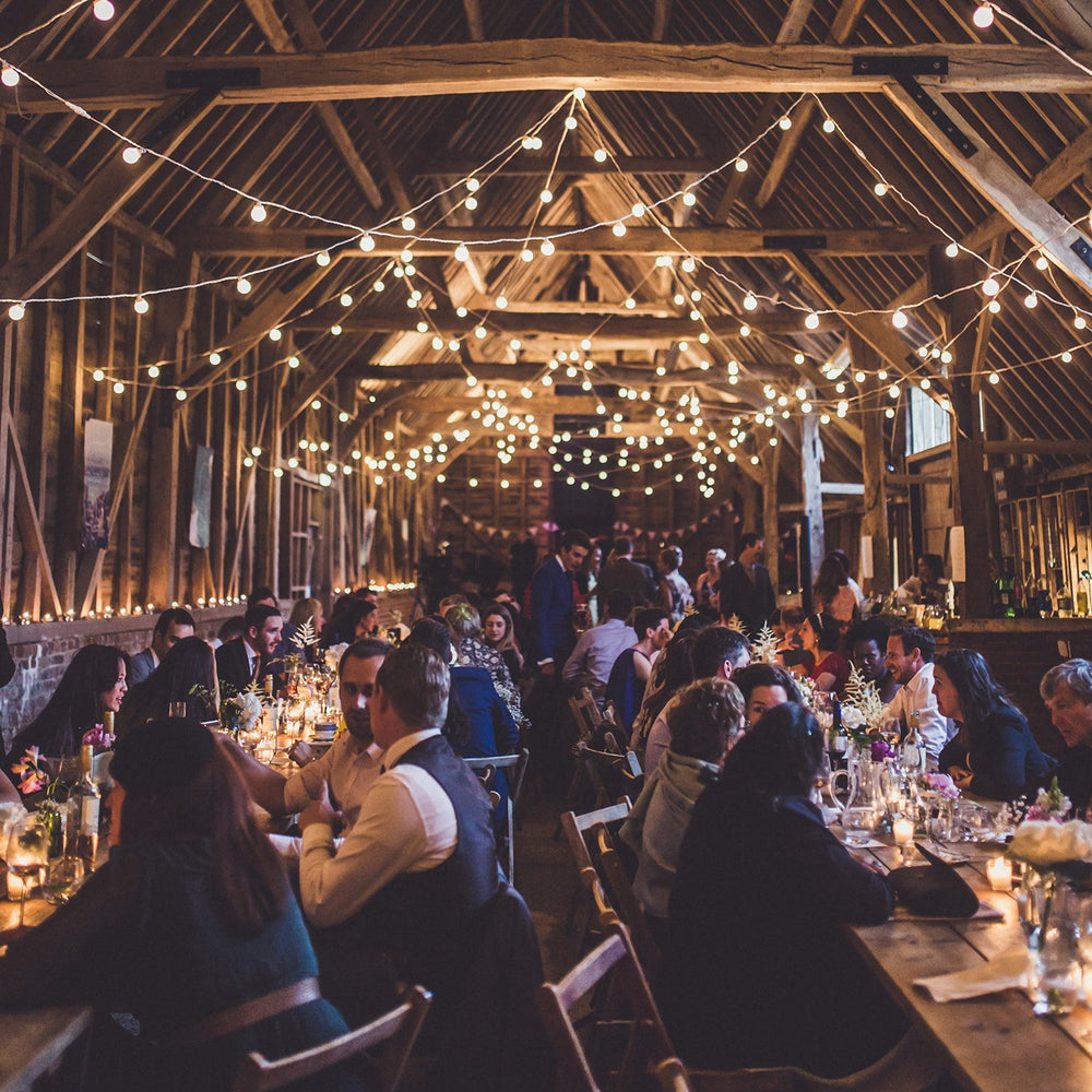 Hochzeitsparty in Scheune mit mehreren warmweißen Lichterketten über den Tischen