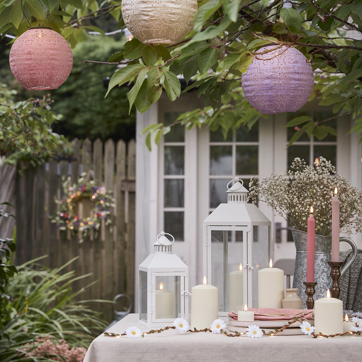 Pastellfarbene Solar Lampions hängen über einem gedeckten Gartentisch mit Kerzen und Blumen