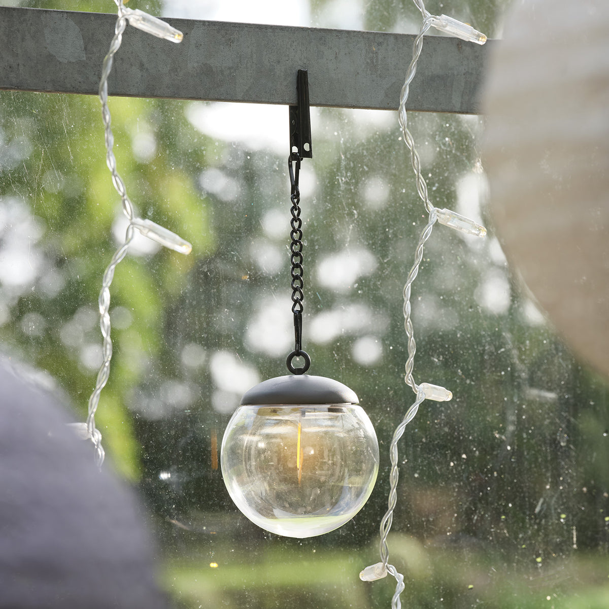 Transparente Solar-Hängelaternenkugel mit Filament-LED hängt an Clip vor sonniger, nasser Fensterscheibe