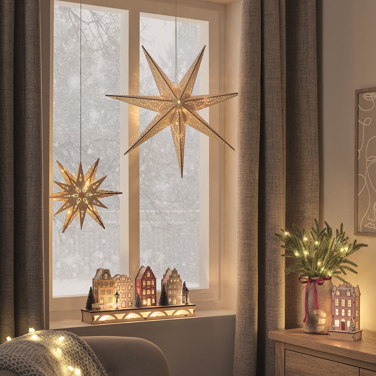 Beleuchtetes LED-Winterdorf auf Fensterbank mit zwei hängenden Papiersternen und Weihnachtsdeko