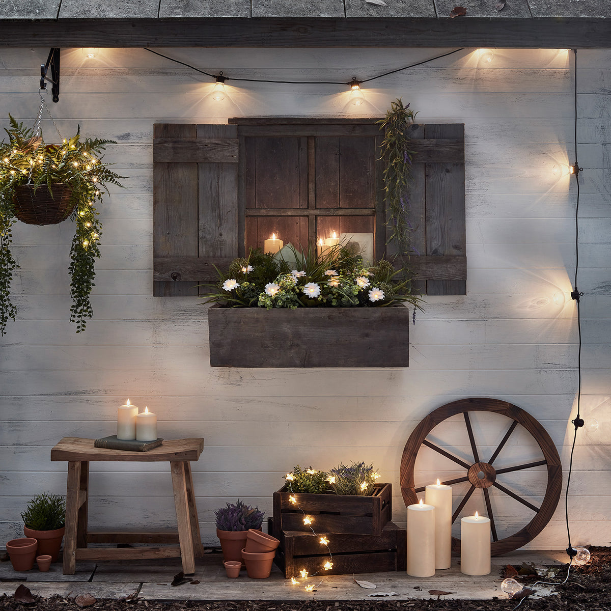Lichterkette mit warmweißen Glühbirnen an weißer Holzfassade mit Kerzen, Pflanzen und Holzhocker