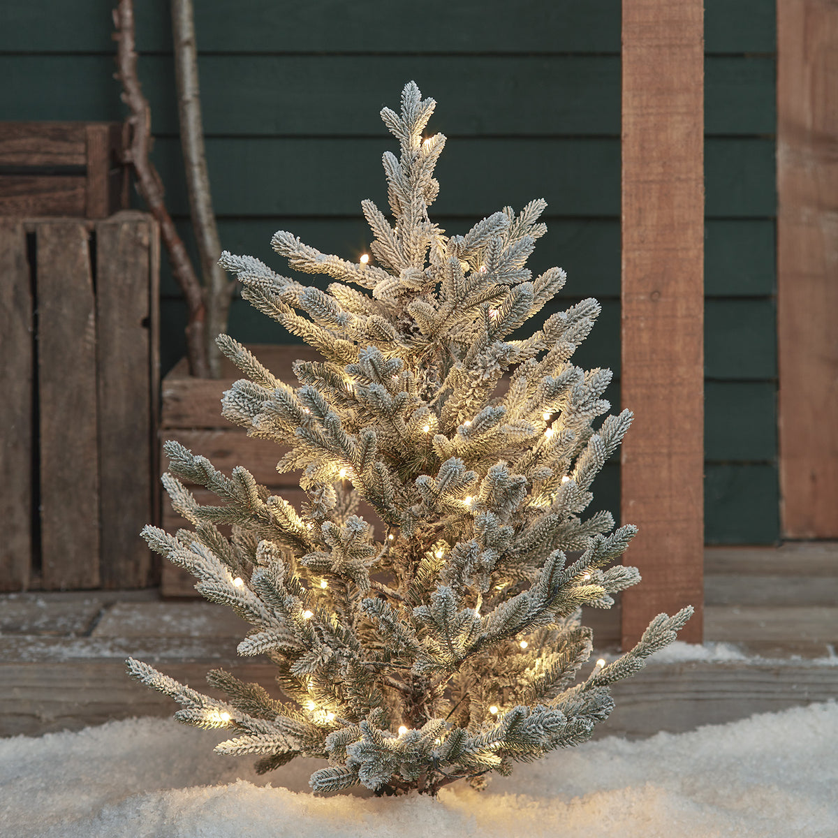 Verschneiter Weihnachtsbaum mit warmweißen LED-Lichtern im Schnee vor Holzverkleidung