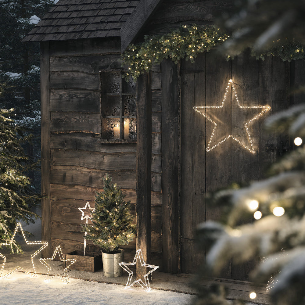Beleuchteter Weihnachtsbaum im Topf vor Holzhütte mit leuchtenden Sternen und Schnee