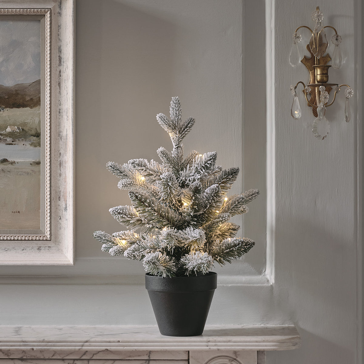 Verschneiter Mini-Tannenbaum mit warmweißen LEDs in schwarzem Topf auf weißem Kaminsims
