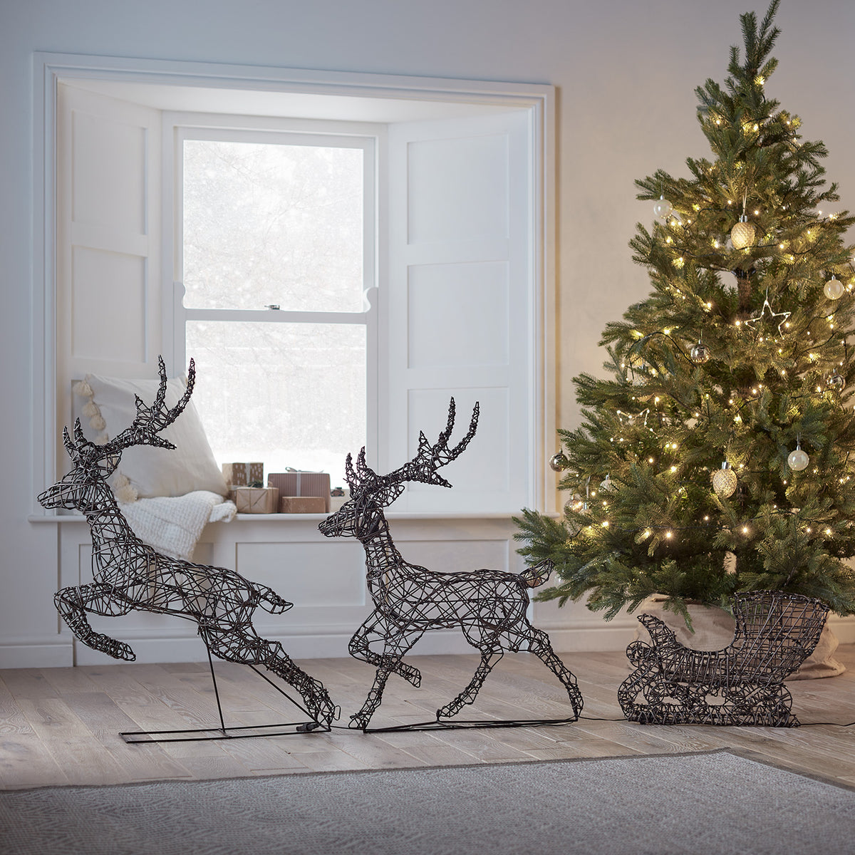Zwei unbeleuchtete Rentiere und Schlitten in Rattan-Optik vor Fenster mit Weihnachtsbaum