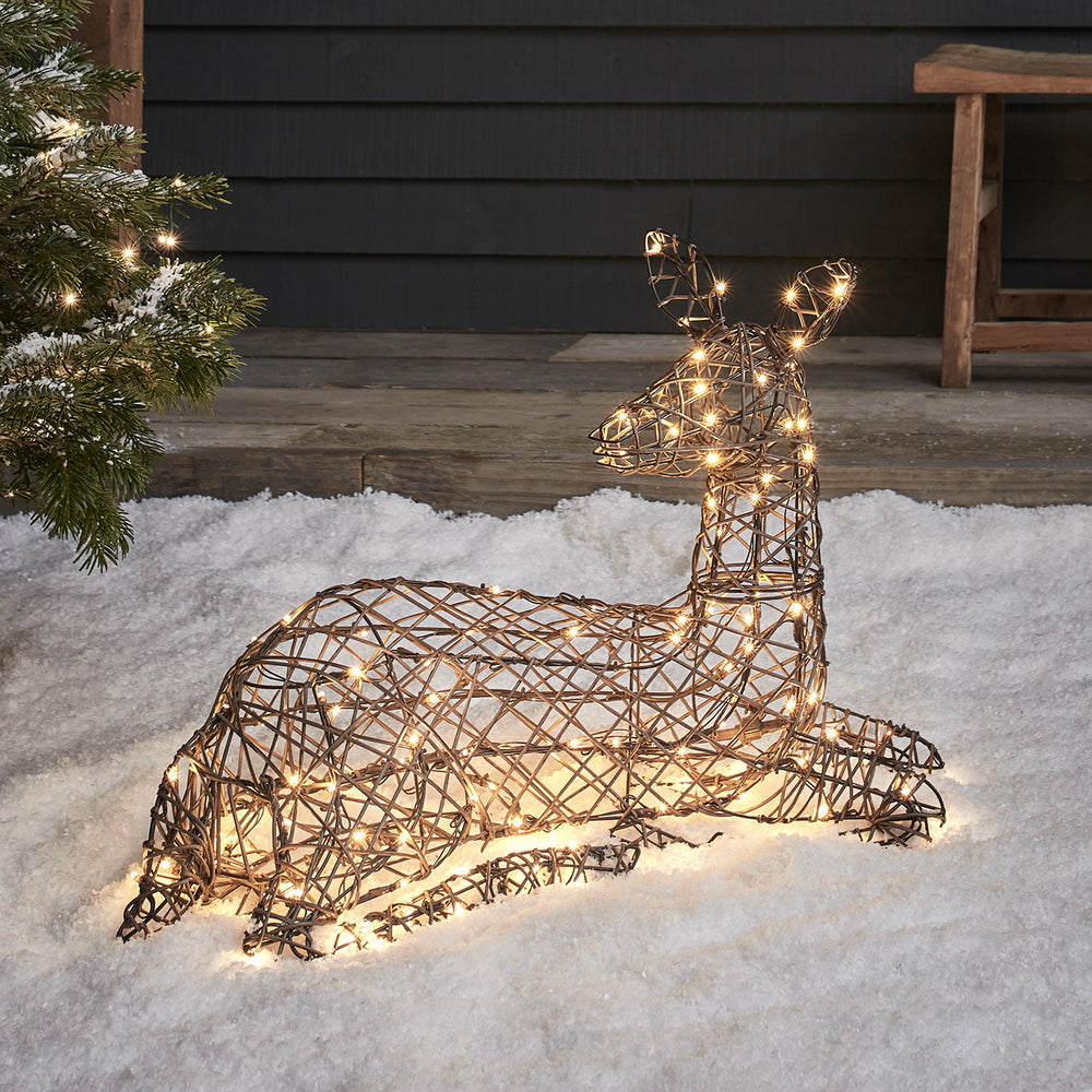 Solar-LED-Rentier in brauner Rattan-Optik liegt beleuchtet im Schnee vor dunkler Hauswand