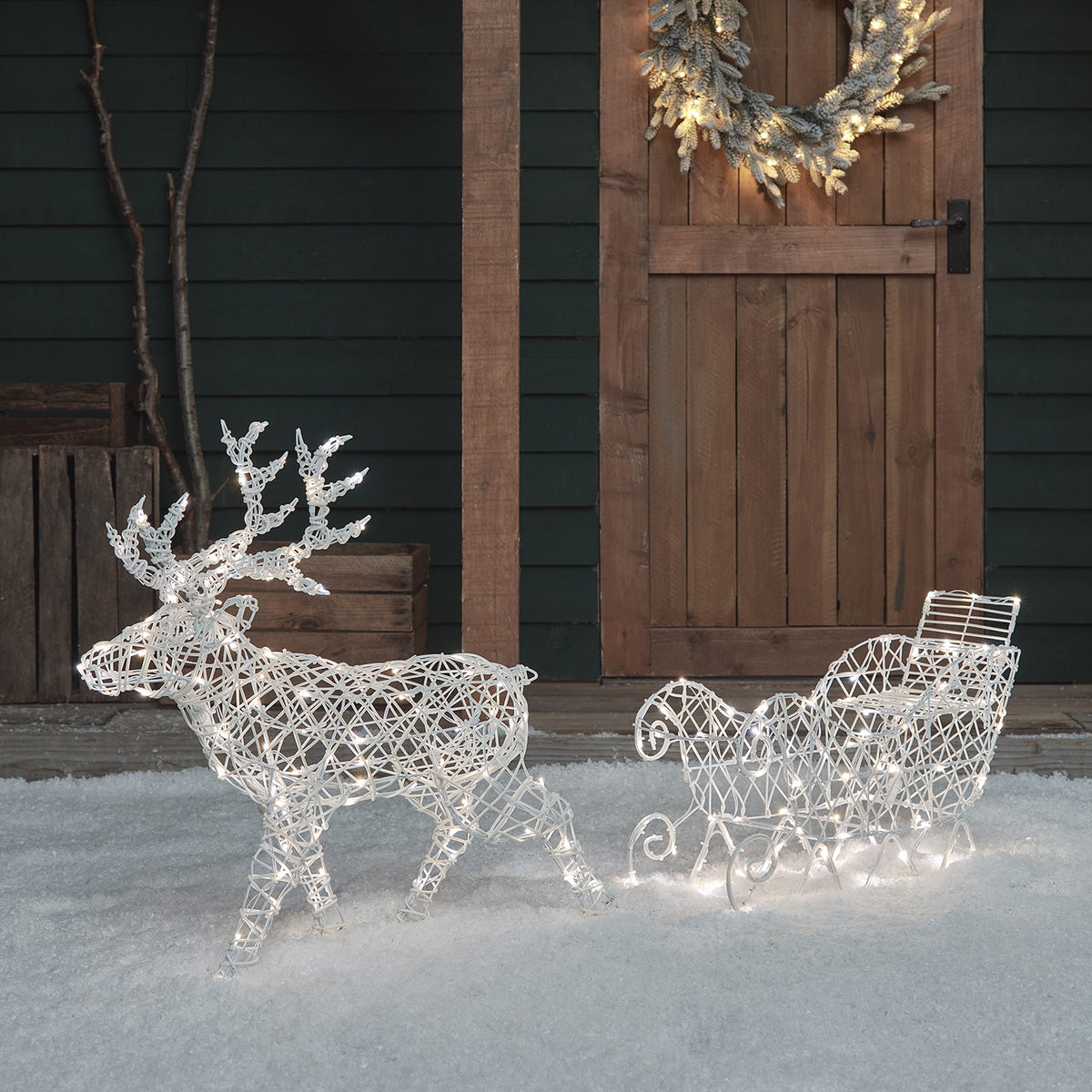LED-Rentier mit Schlitten beleuchtet mit warmweißem Licht auf Schneefläche vor Holztür