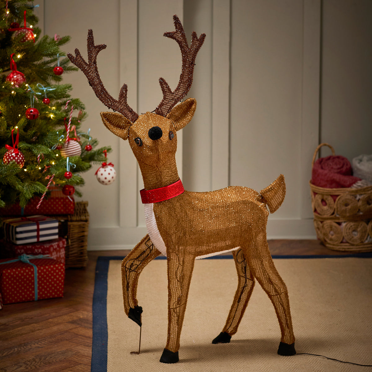 Nahaufnahme des beleuchteten LED-Rentiers mit rotem Schal vor Weihnachtsbaum und Körben