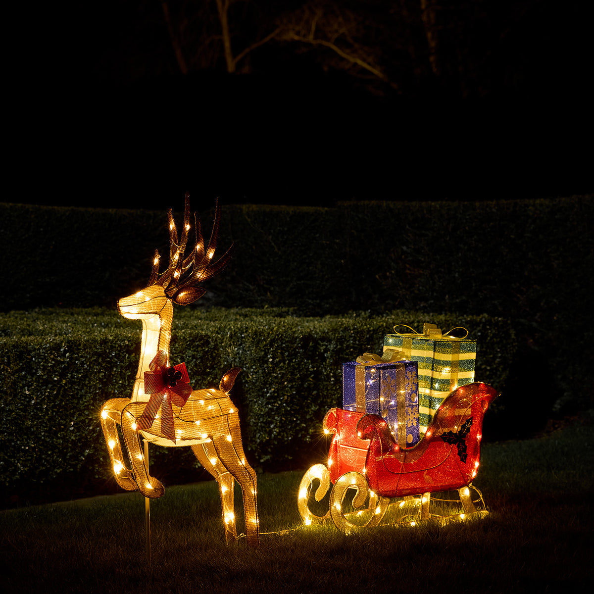 Leuchtendes LED-Rentier mit roter Schleife zieht roten Schlitten mit bunten Geschenkboxen im Garten bei Nacht
