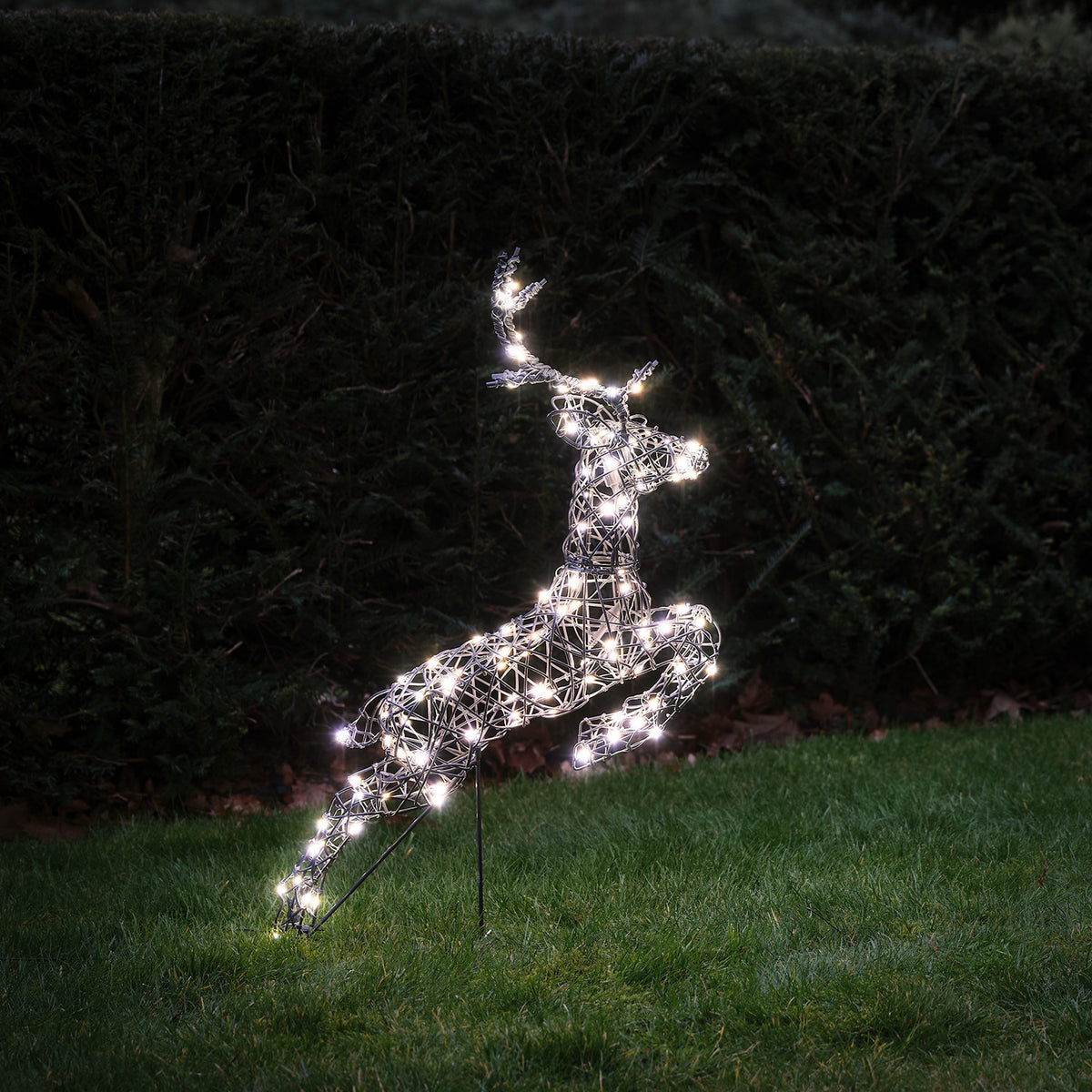Springendes LED-Rentier mit warmweißen Lichtern vor roter Holzwand und Tannenzweigen im Schnee