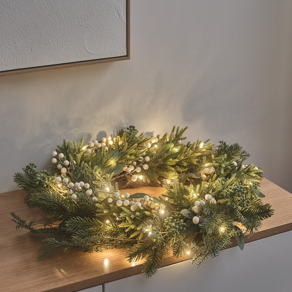 Weihnachtskranz mit künstlichem Tannengrün, weißen Beeren und warmweißen LEDs auf Holzfläche