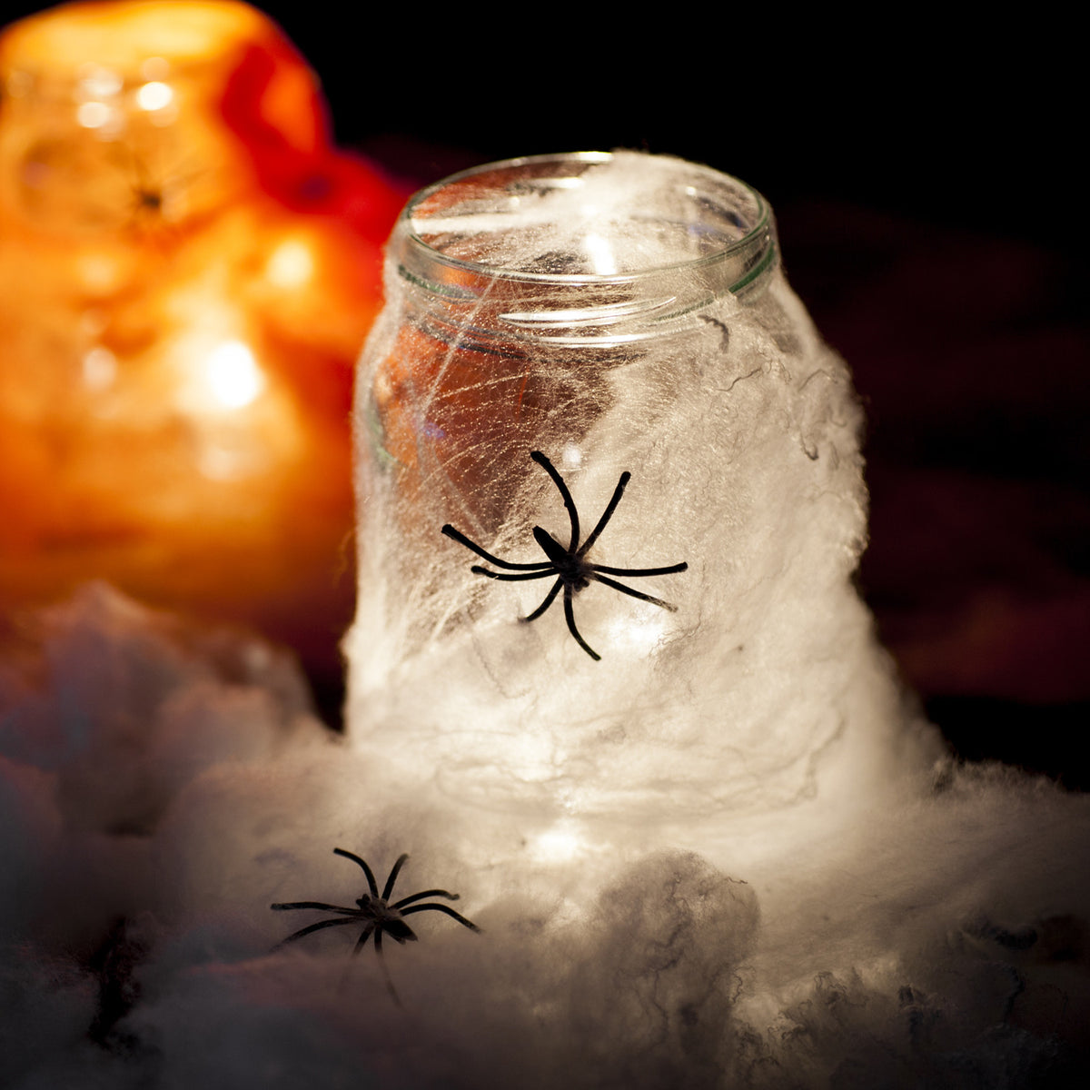 Glas mit weißem Spinnennetz und schwarzen Spinnen, im Hintergrund orangefarbene Dekoration