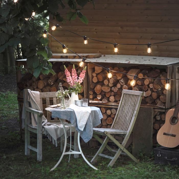 Lichterkette mit warmweißen Glühbirnen über Gartentisch mit Stühlen und Holzstapel