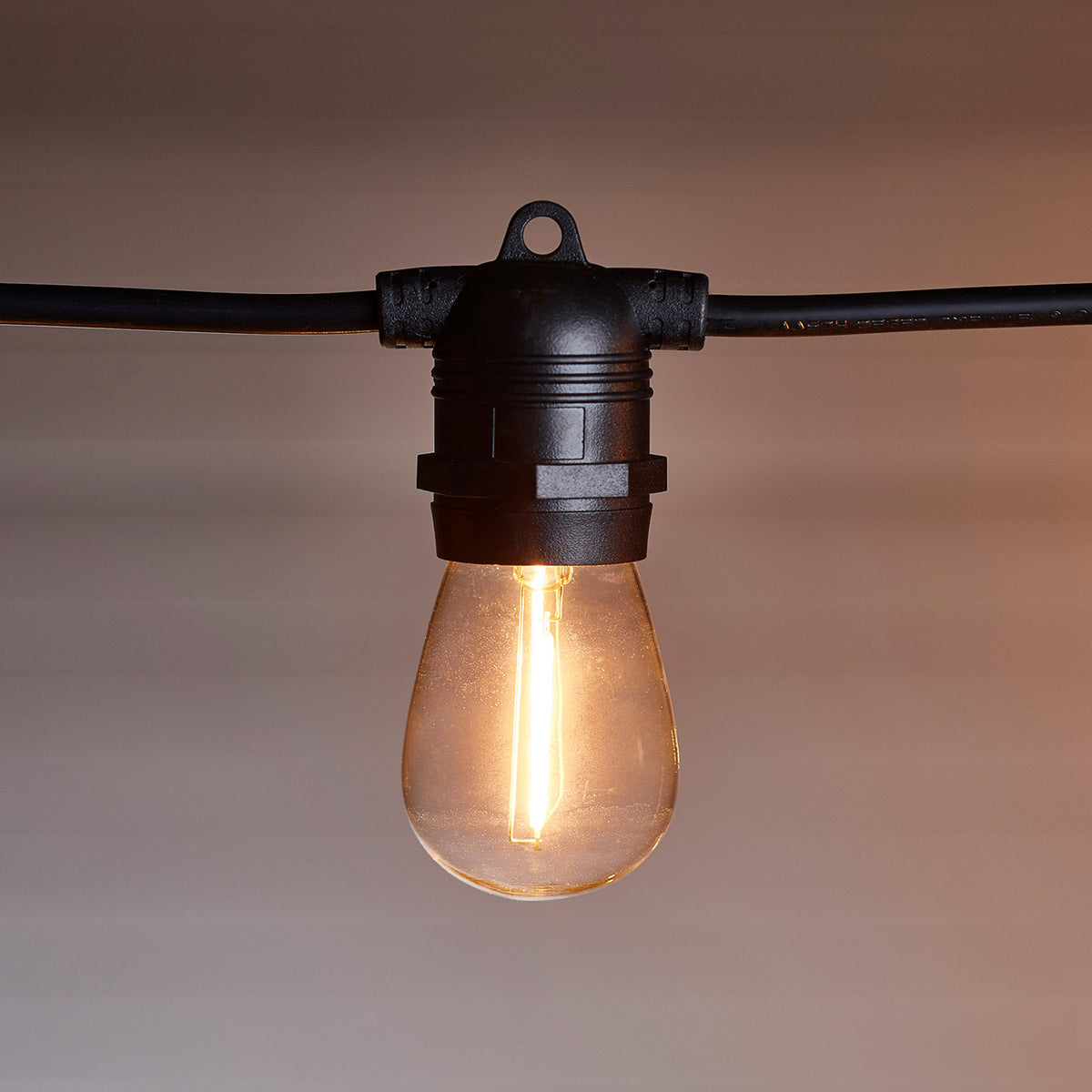 Warmweiß leuchtende Glühbirne an schwarzem Kabel mit unscharfem Hintergrund im Garten