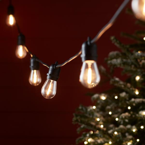 Lichterkette mit warmweißen Glühbirnen vor rotem Hintergrund und Weihnachtsbaum