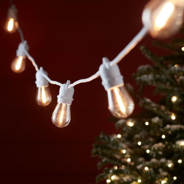 Warmweiße Glühbirnen an weißem Kabel vor rotem Hintergrund mit Weihnachtsbaum im Hintergrund