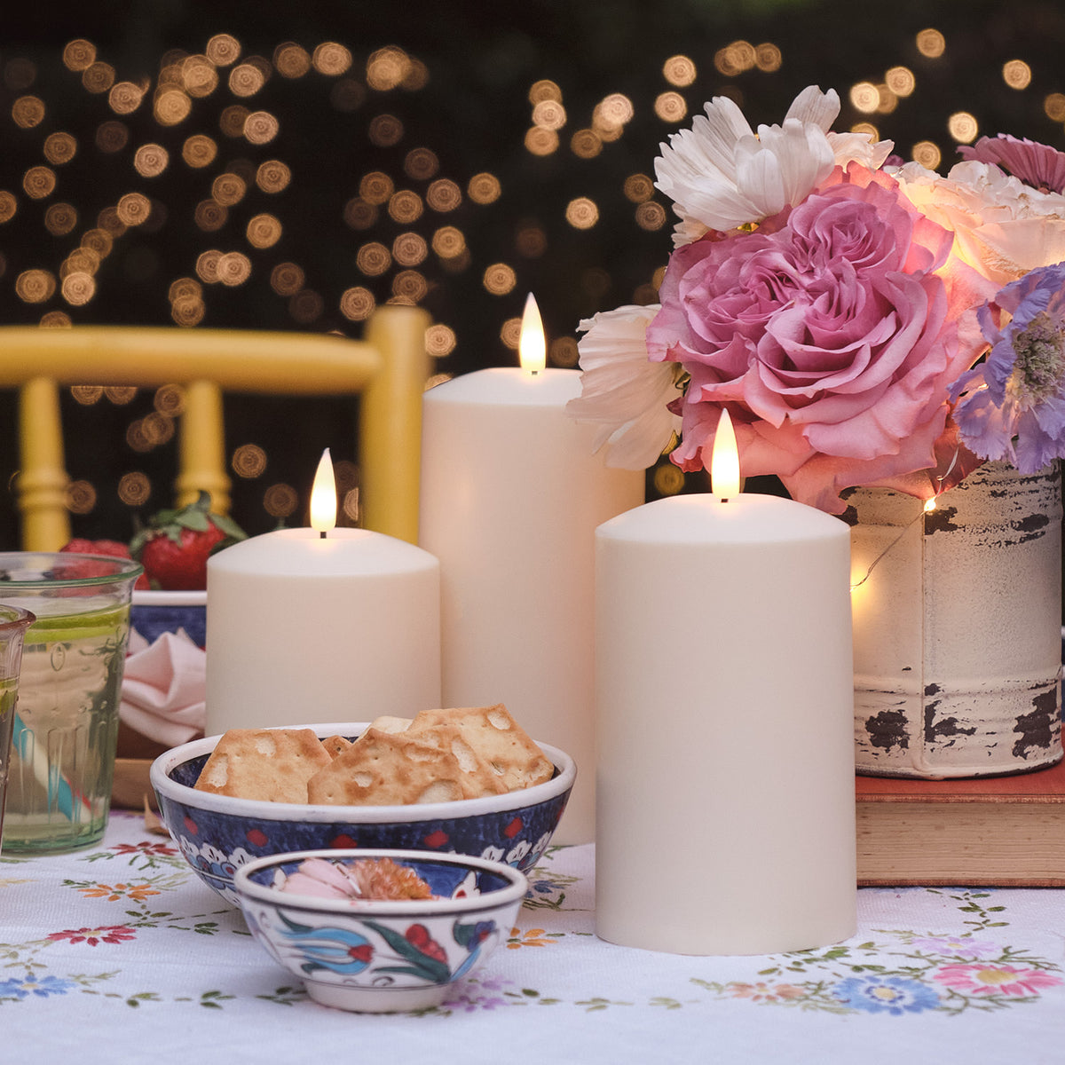 Drei TruGlow LED Kerzen auf gedecktem Gartentisch mit Blumenarrangement und Snacks