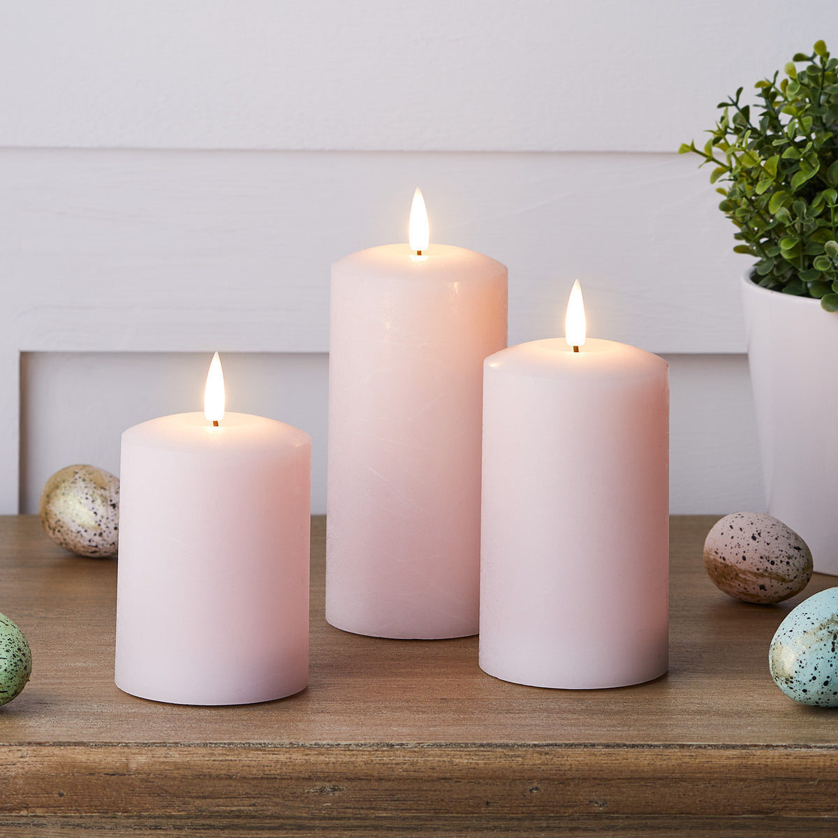 Drei rosa LED Stumpenkerzen mit Osterdeko und bemalten Eiern auf Holztisch