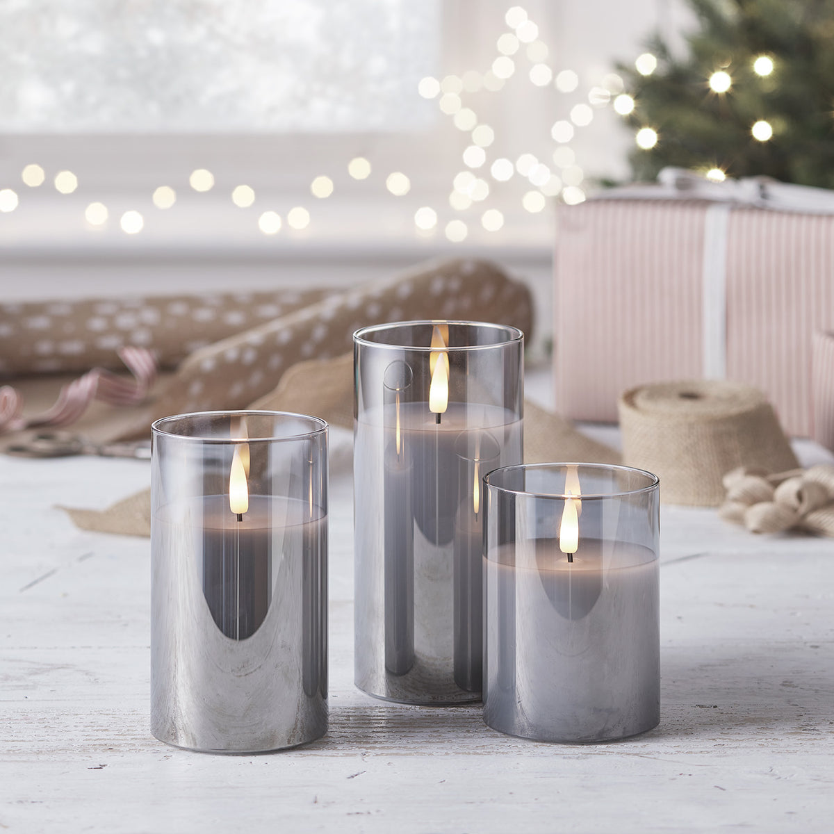 Trio TruGlow LED Kerzen in rauchfarbenen Gläsern dekorativ auf Tisch mit Weihnachtsdeko im Hintergrund