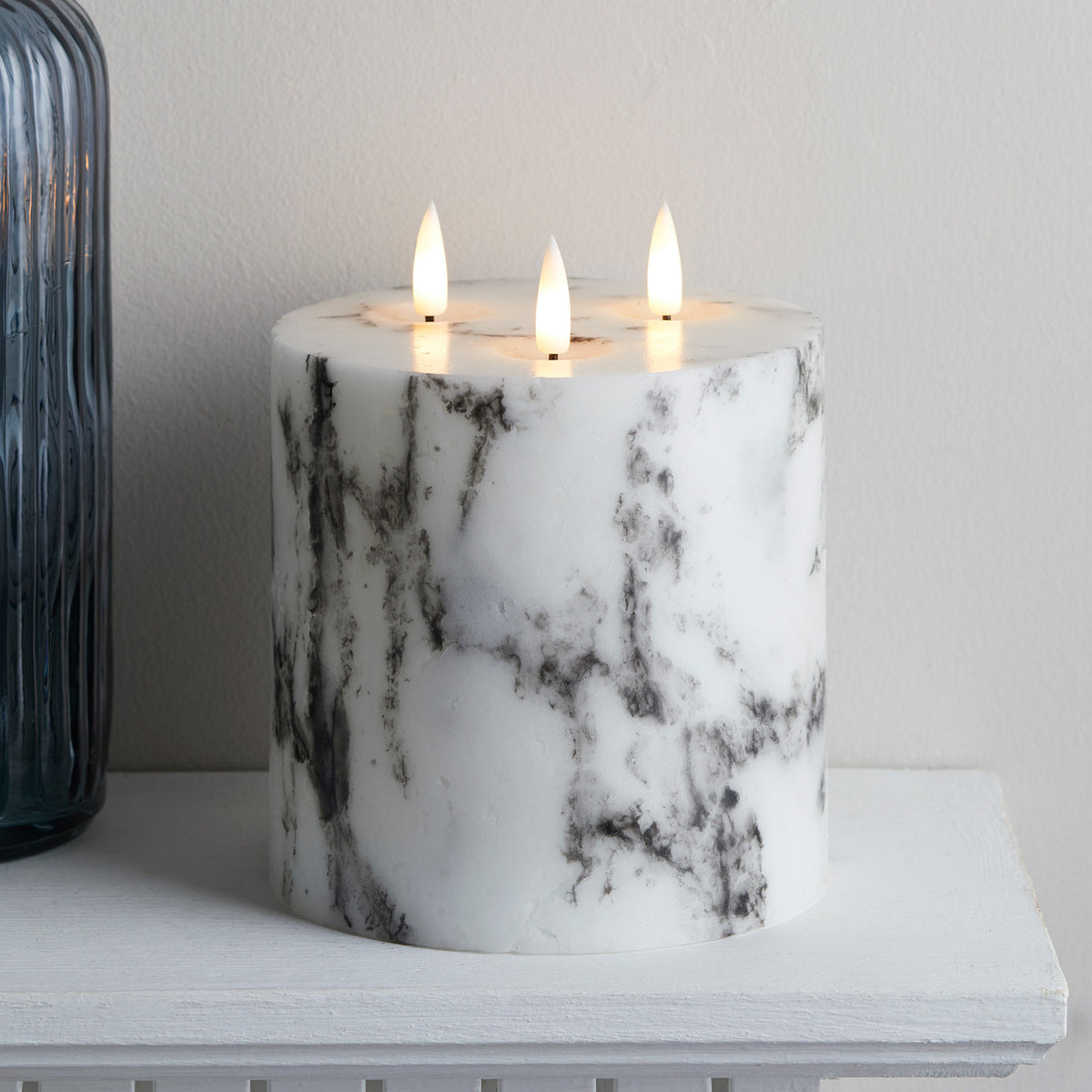 Nahaufnahme der dreidochtigen LED Kerze mit Marmoroptik auf weißem Regal neben blauem Glasvasen