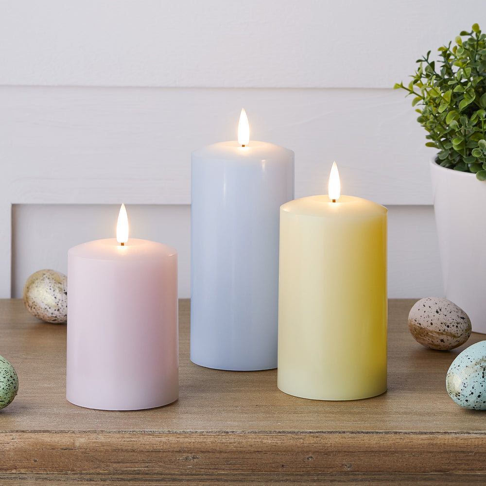 Drei pastellfarbene LED Stumpenkerzen in Rosa, Blau und Gelb auf Holztisch mit Ostereiern