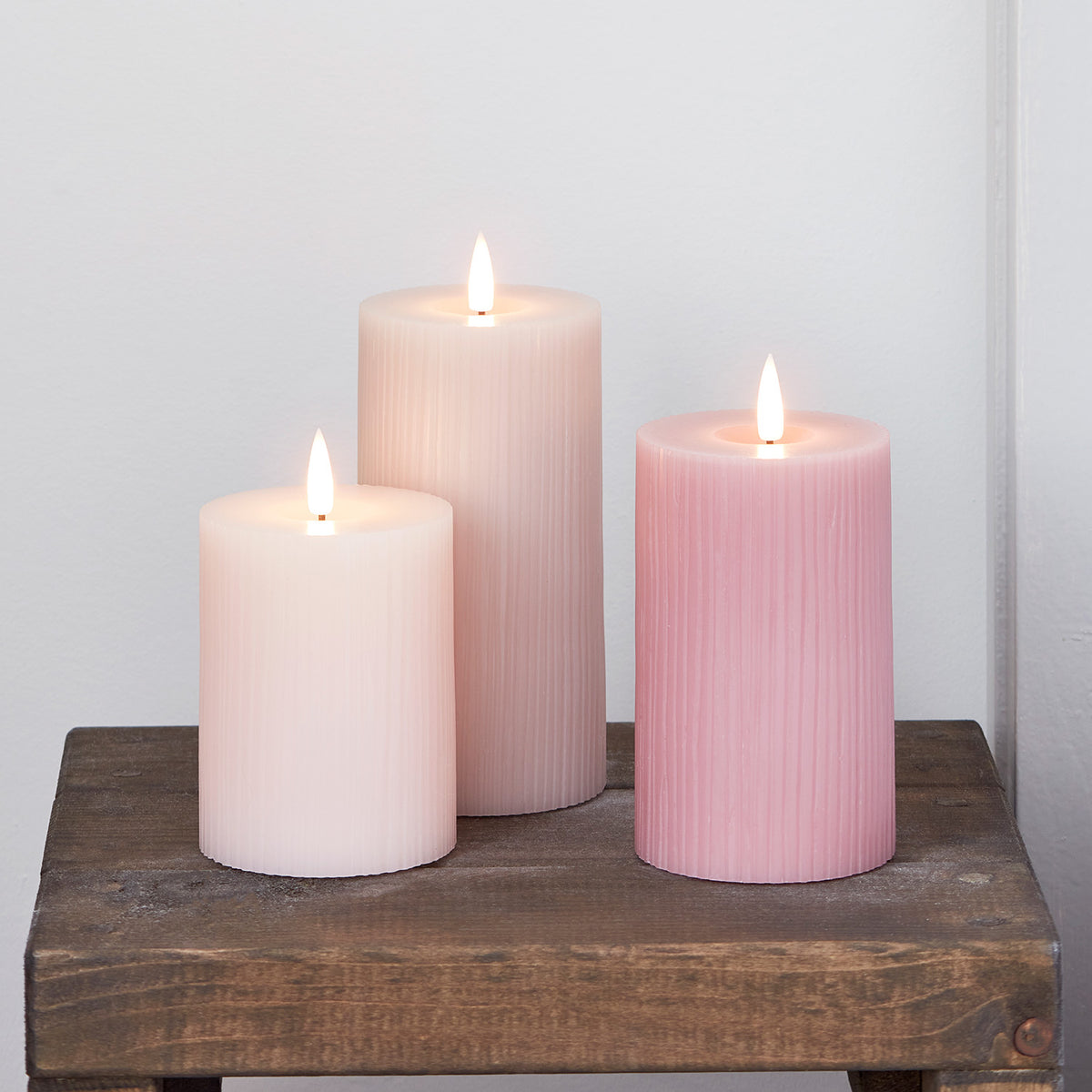 Drei LED-Kerzen in Rosa- und Pastelltönen auf dunklem Holztisch vor weißer Wand
