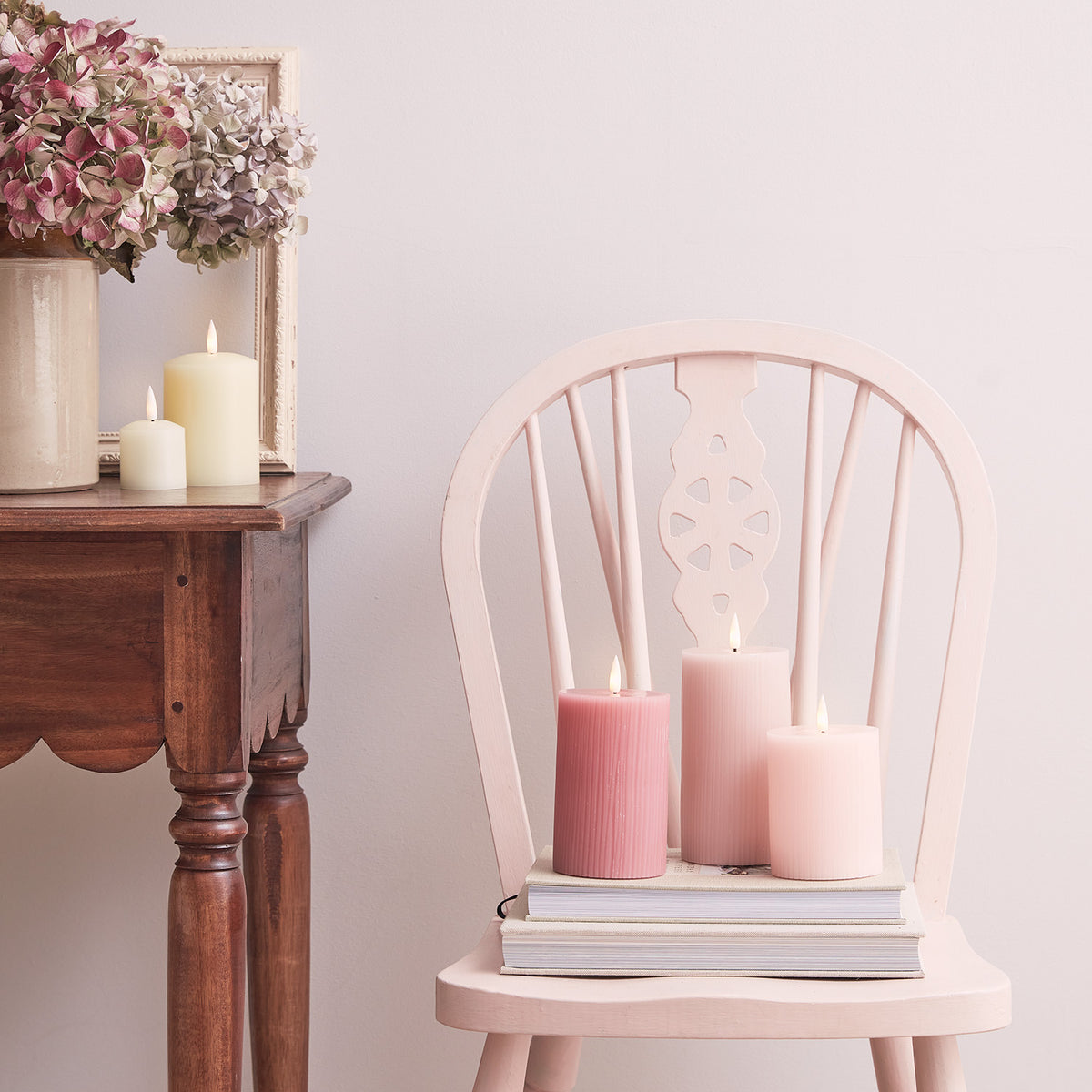 Drei brennende LED-Kerzen in Rosa- und Pastelltönen auf einem rosa Stuhl mit Büchern