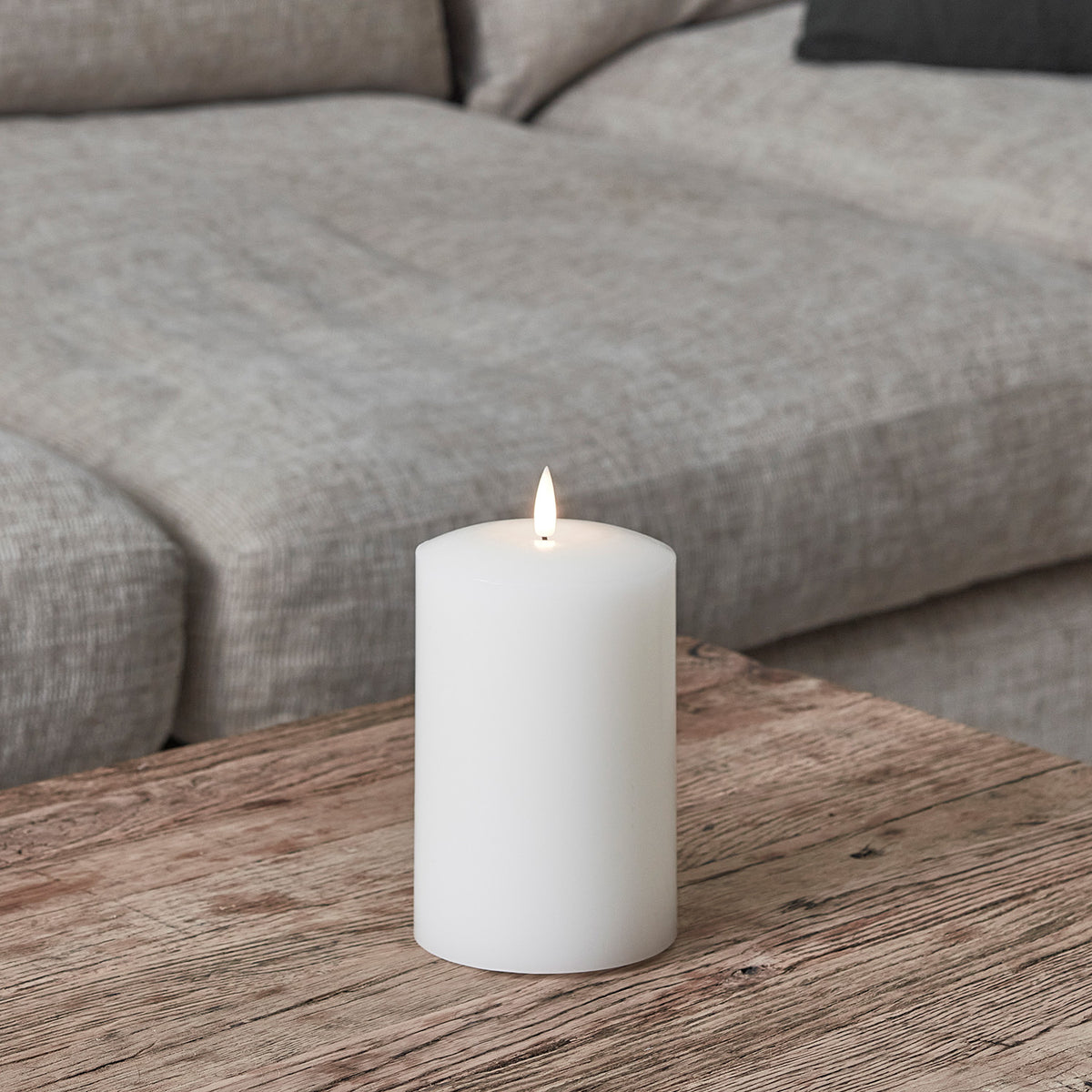 Weiße breite LED Stumpenkerze aus Echtwachs auf Holztisch vor grauem Sofa
