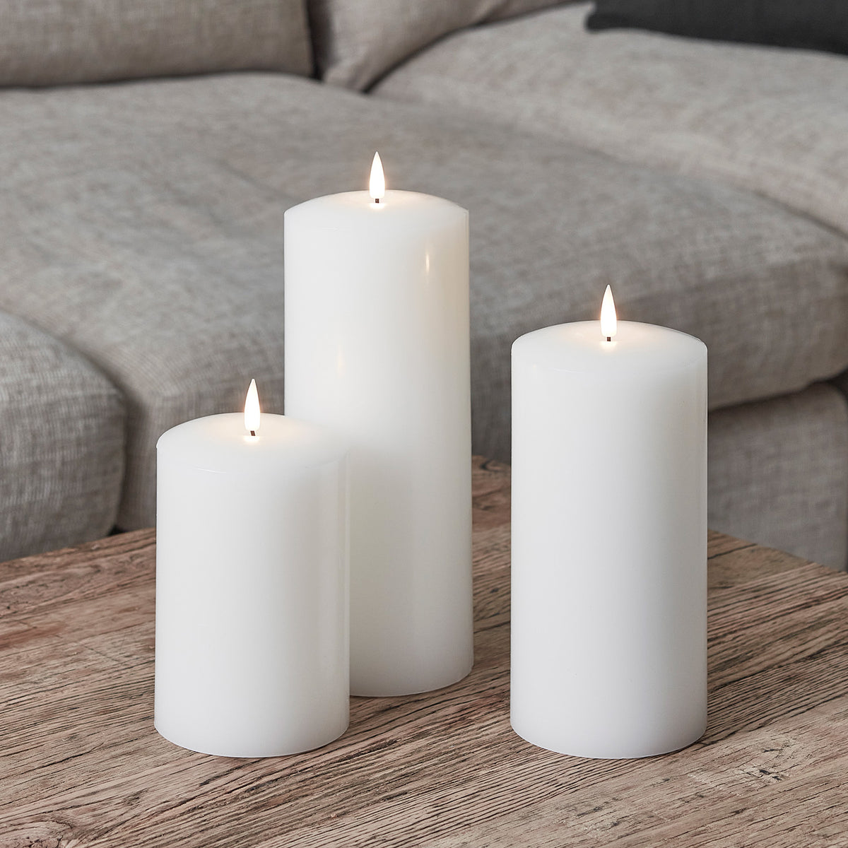 Drei weiße LED Echtwachs-Kerzen in verschiedenen Höhen auf Holztisch vor grauem Sofa