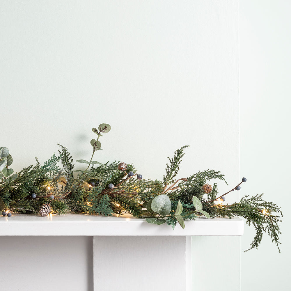 Weihnachtsgirlande mit Tannenzweigen, Eukalyptus, Beeren, Tannenzapfen und warmweißen Lichtern auf Kaminsims