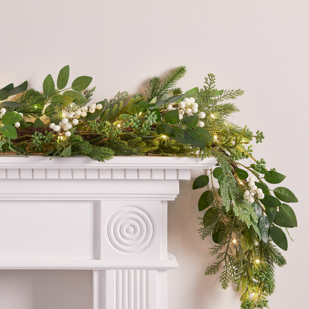 Nahaufnahme der grünen Weihnachtsgirlande mit weißen Beeren und warmweißen LED-Lichtern auf weißem Kaminsims