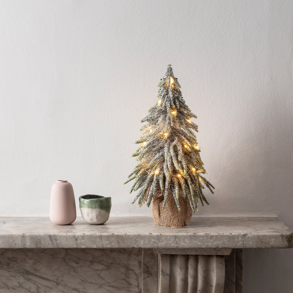 Mini Weihnachtsbaum mit Schnee und warmweißer Micro Lichterkette auf Kaminsims neben zwei Vasen