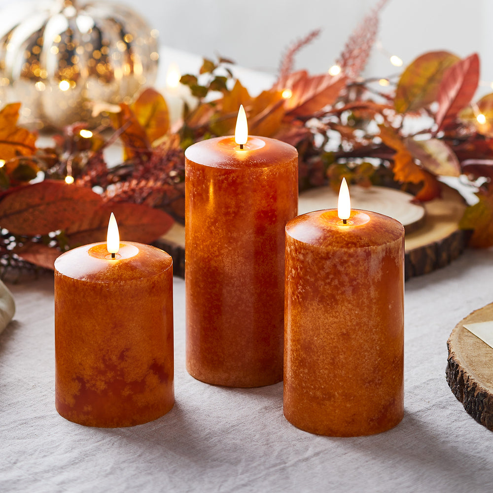 Drei unterschiedlich hohe orangefarbene LED-Kerzen mit flackernden Flammen vor herbstlicher Dekoration