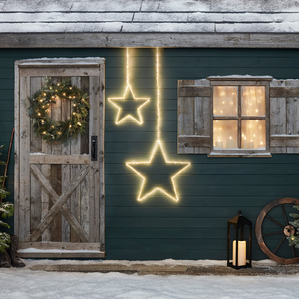 Zwei leuchtende LED-Sterne an einer grünen Hauswand mit Tür, Fenster und winterlicher Dekoration