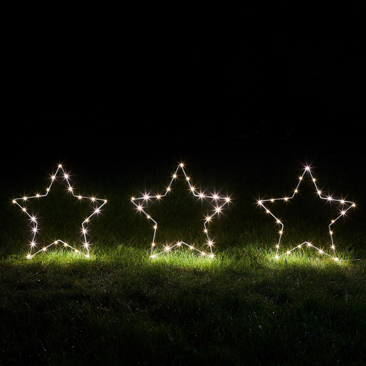 Drei leuchtende Micro LED-Sterne nebeneinander auf Gras bei Dunkelheit