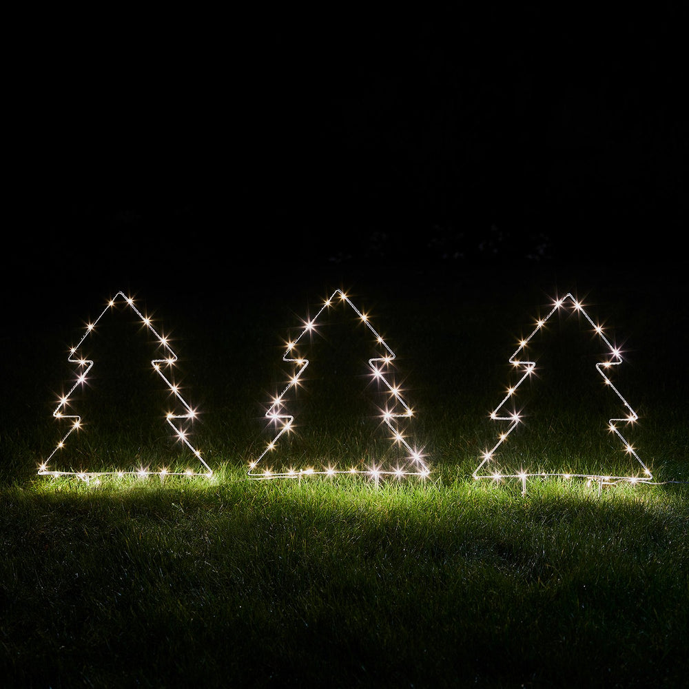 Drei leuchtende Micro LED Tannenbäume aus Metallrahmen nebeneinander auf Gras bei Nacht