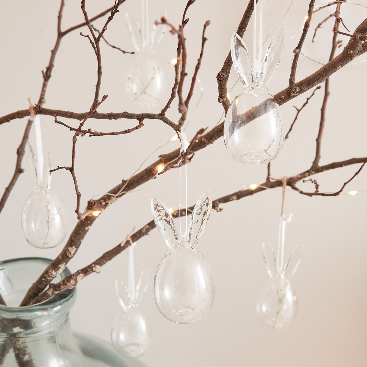 Transparente Ostereier mit Hasenohren hängen an Zweigen in einer Glasvase mit Lichterkette
