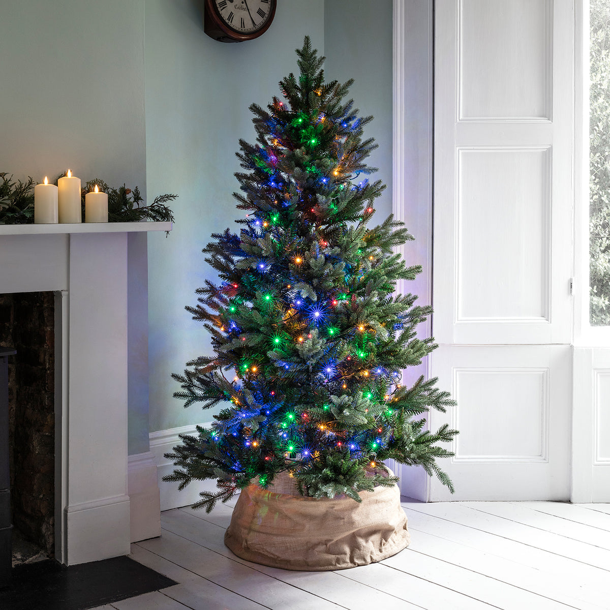 Weihnachtsbaum mit bunter LED-Lichterkette in einem hellen Raum neben Kamin und Fenster