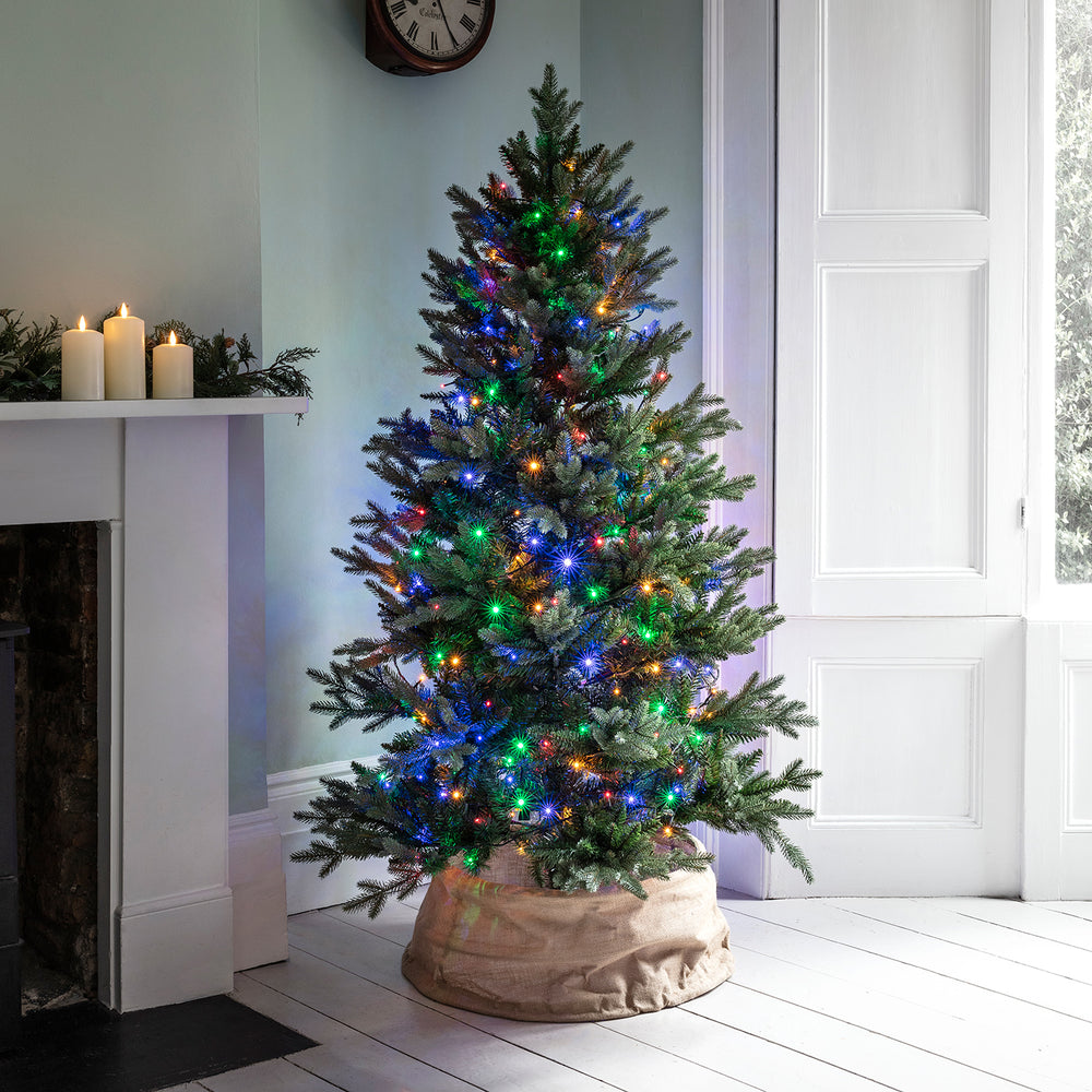 Weihnachtsbaum mit bunter LED Lichterkette in einem hellen Raum neben Kamin und Fenster
