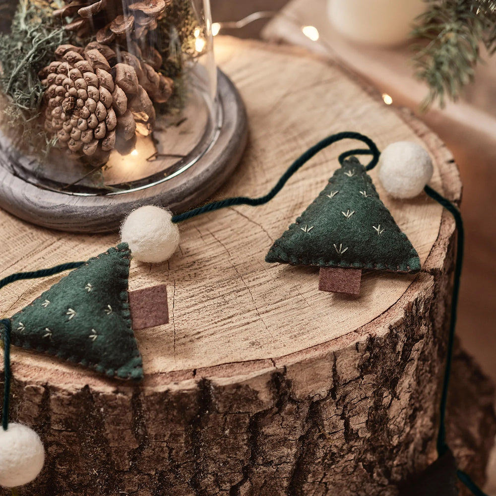 Nahaufnahme der Weihnachtsgirlande mit Filztannenbäumen und Filzkugeln auf rustikalem Holzstamm neben Glasglocke