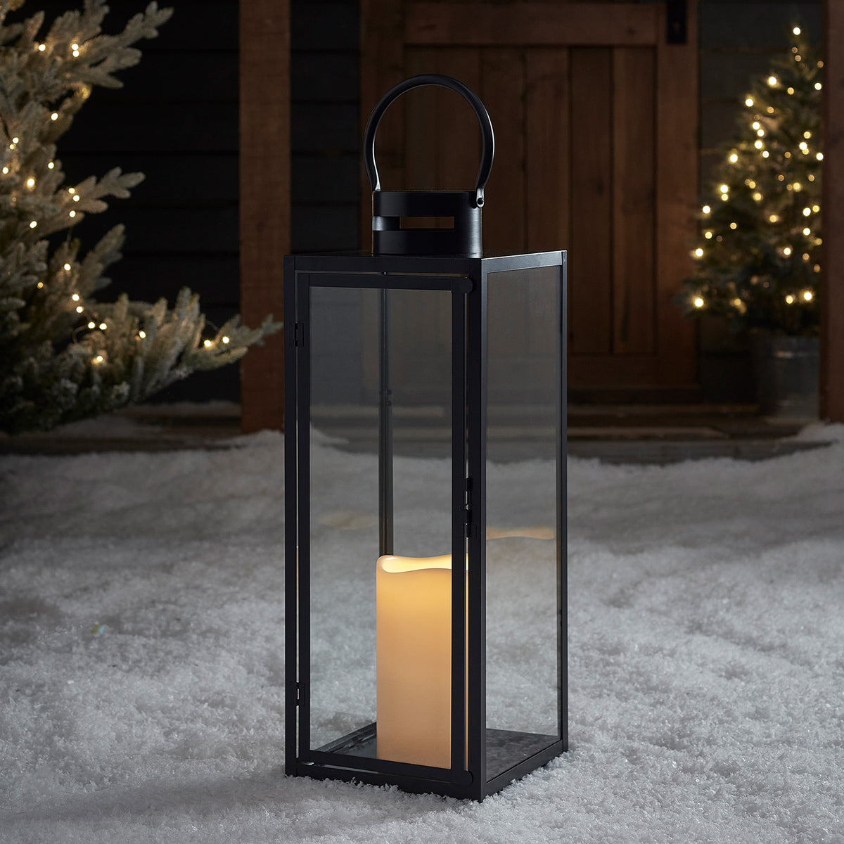 Große dunkelgraue Metall-Laterne mit Glasfenstern und warmweiß flackernder LED-Kerze im Schnee vor Holztür