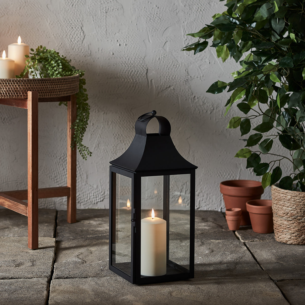 Schwarze Gartenlaterne mit TruGlow Outdoor Kerze auf Steinboden neben Pflanzen und Holzstuhl