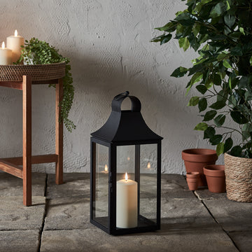 Schwarze Gartenlaterne mit TruGlow Outdoor Kerze auf Steinboden neben Pflanzen und Holzstuhl