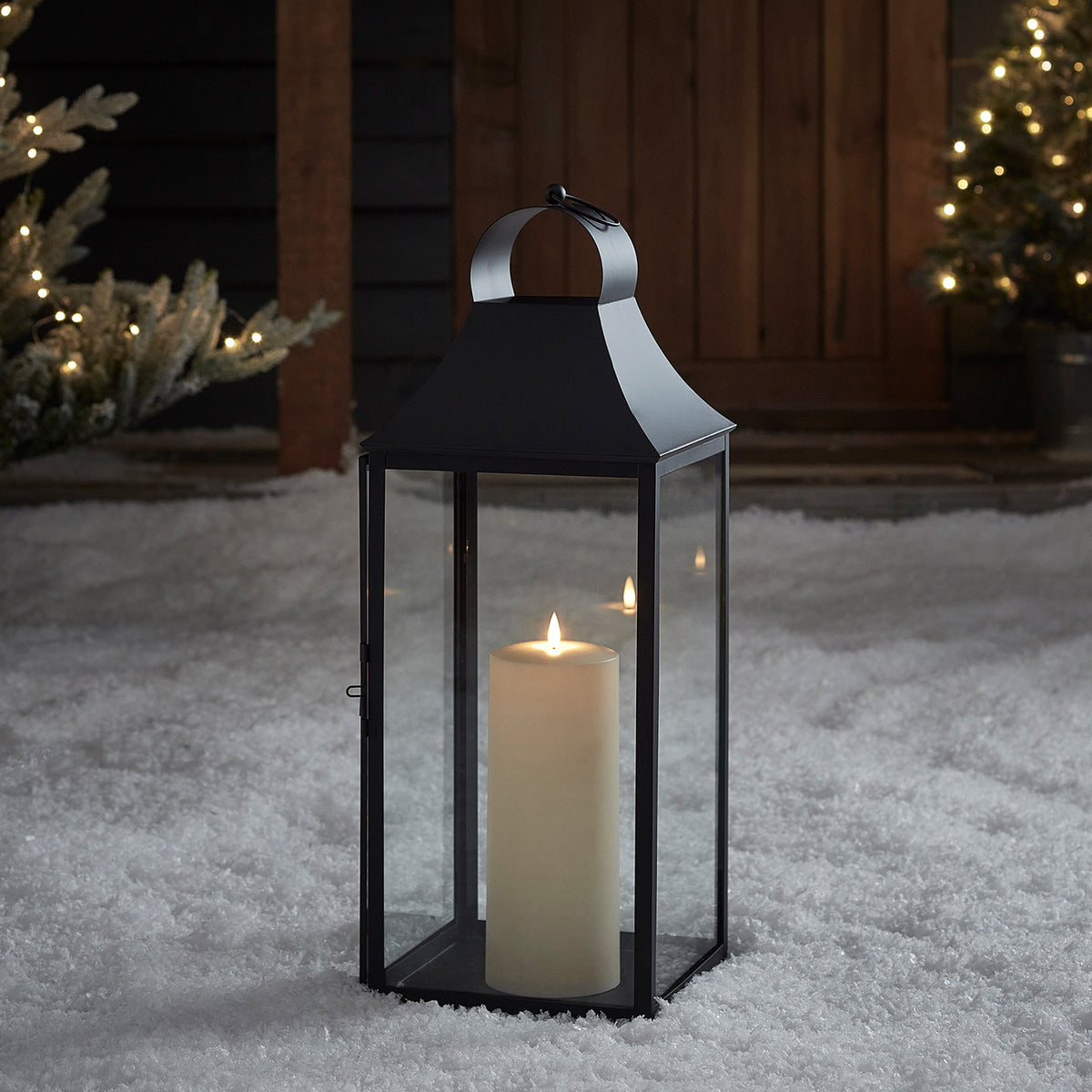 Schwarze Gartenlaterne mit TruGlow Outdoor Kerze leuchtet im Schnee vor Holztür und Lichterkette