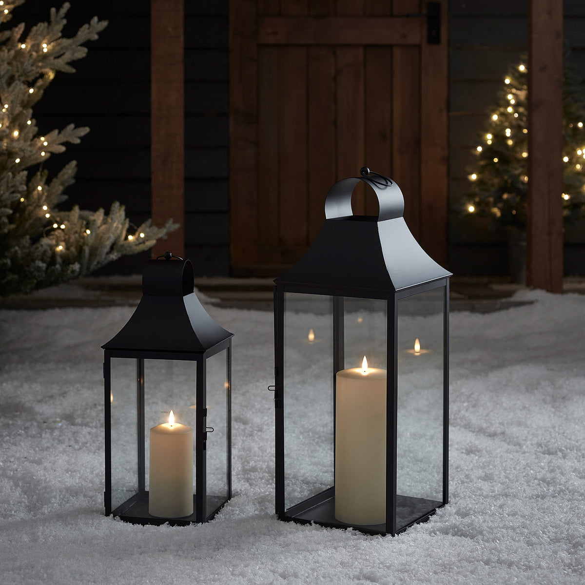 Zwei schwarze Laternen mit LED-Kerzen im Schnee vor Holztor und beleuchteten Bäumen