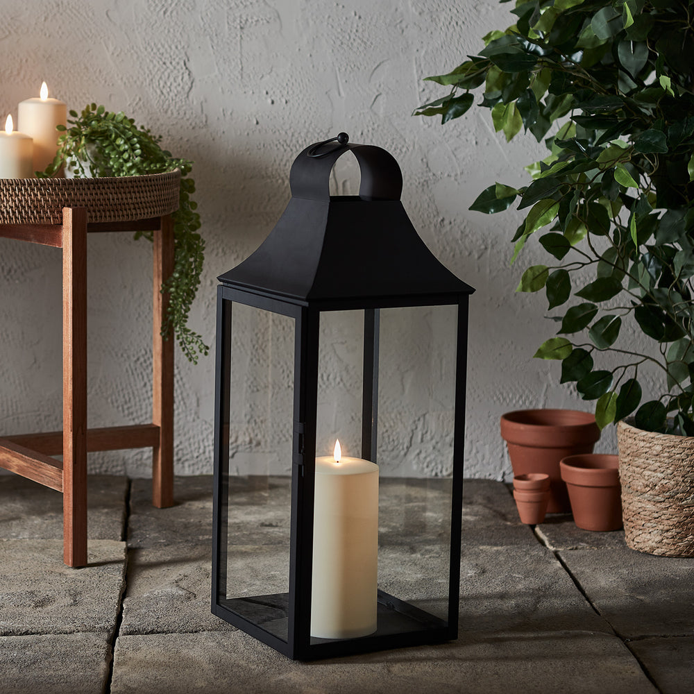 Schwarze Gartenlaterne mit TruGlow Outdoor LED-Kerze auf Steinboden neben Pflanzen und Holzstuhl