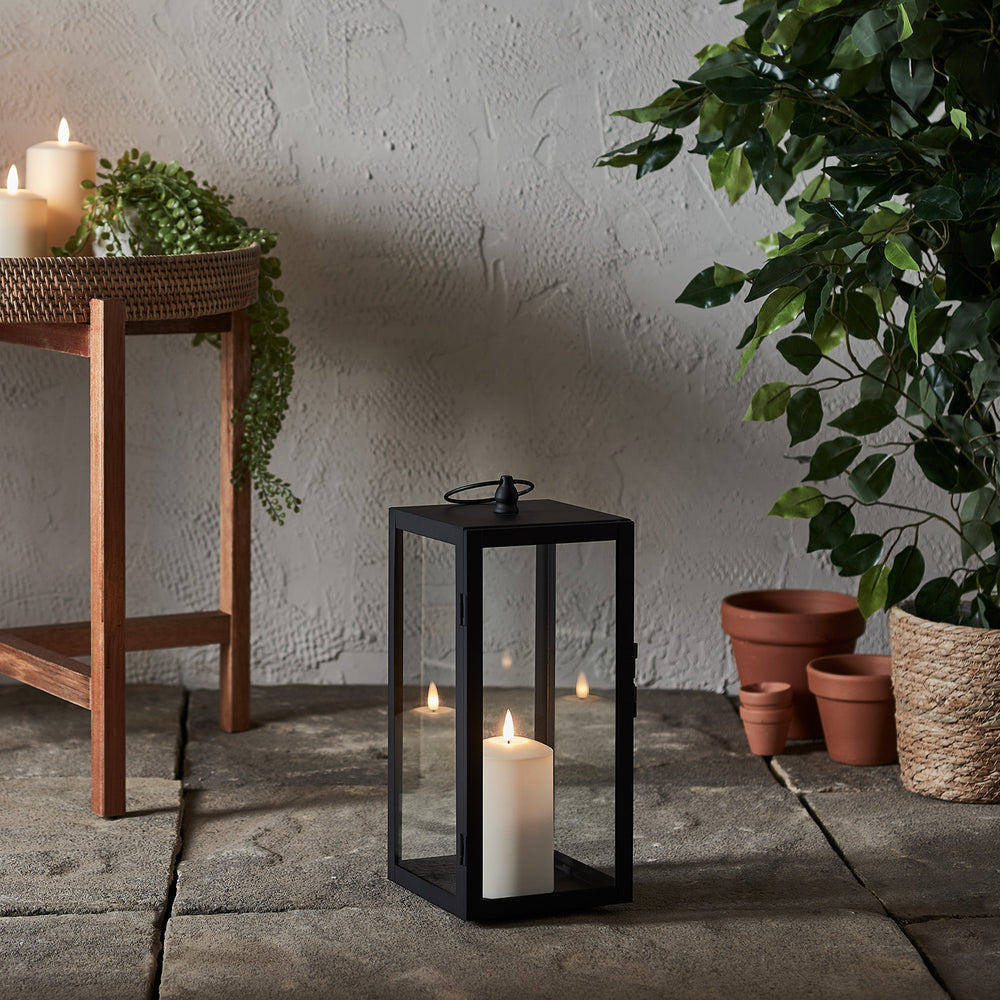 Schwarze Gartenlaterne mit TruGlow Outdoor Kerze auf Steinboden neben Holztisch und Pflanzen