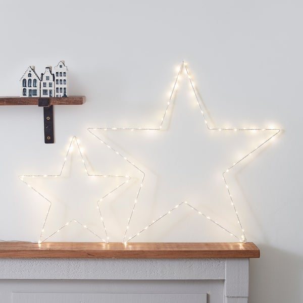 Großer und kleiner weißer LED-Stern mit warmweißen LEDs auf Holztisch vor weißer Wand mit Deko
